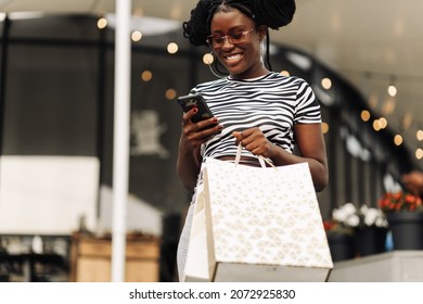 Beautiful fashionable african american girl holding bags, using smartphone and smiling while shopping at womens boutique for Black Friday, shopping concept, black friday sale