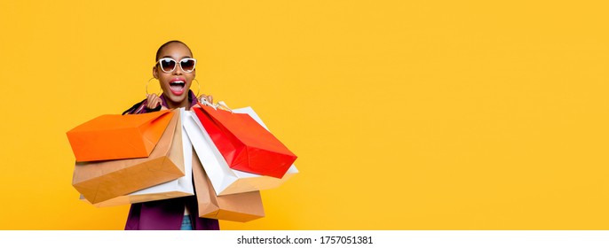 Beautiful fashionable African American woman with colorful shopping bags in surprised sale concept isolated on yellow banner background