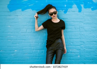 Beautiful fashionable asian girl wearing sunglasses and black clothes outfit posing in front of blue wall
