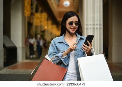 Beautiful fashionable asian woman carrying shopping bags and using smart phone outdoor front of the shopping center - Powered by Shutterstock