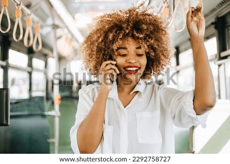 Beautiful and fashionable black woman standing in city bus. She is happy and smiled while using her smartphone to communicate with someone. Modern city lifestyle concept. Bright sunny day.