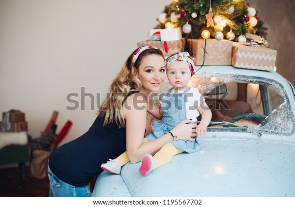 Beautiful and fashionable blonde mom with little\
cute daughter posing together, sitting on blue retro car and\
looking at camera. Stylish parent and female kid in stylish\
decorated for Christmas\
studio