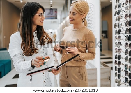Beautiful and fashionable blonde woman choosing eyeglasses frame in modern optical store. Female seller specialist helps her to make right decision.
