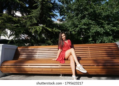 Beautiful Fashionable Brunette Woman In Red Dress Sits On A City Bench