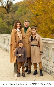 Beautiful Fashionable Family: Mother, Father, Two Children (sons) Are Posing, Standing In Autumn Park,yellow Orange Leaves Trees. People Wear Warm Beige Brown Coats,boots,trousers. Lifestyle. Vertical