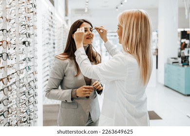Beautiful and fashionable woman choosing eyeglasses frame in modern optical store. Female seller specialist helps her to make right decision.