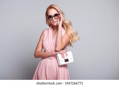 Beautiful fashionable young blonde woman wearing sunglasses, pink dress, purse in a studio. Spring, summer fashion photo over grey; grey background.