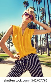 Beautiful Fashionable Young Woman Posing In The Park, Sunglasses, Yellow Top, High Heels, Short Blonde Hair. Fashion Summer Photo. Bright Colors. Nice View