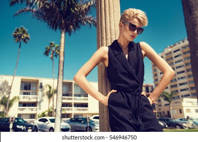 Beautiful Fashionable Young Woman Posing In The Park, Sunglasses, Black Jumpsuit, High Heels, Short Blonde Hair. Fashion Summer Photo. Bright Colors. Nice View