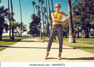 Beautiful Fashionable Young Woman Posing In The Park, Sunglasses, Yellow Top, High Heels, Short Blonde Hair. Fashion Summer Photo. Bright Colors. Nice View