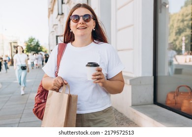 Beautiful fashionable young woman in sunglasses with shopping paper bag standing on city street, black friday. Urban lifestyle concept. Attractive female drinking a coffee in paper bio white cup.