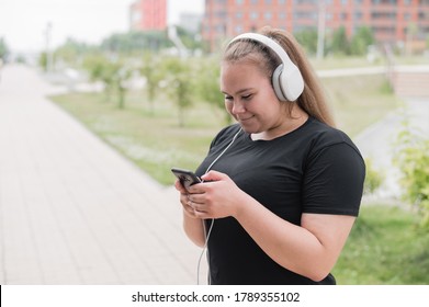 A beautiful fat girl in black sportswear listens to music on headphones and text messages on the phone while walking outdoors. A plump young woman is leafing through a music playlist in smartphone.