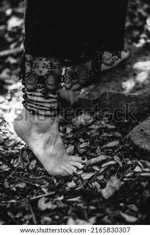 Beautiful feet of young gothic and witch woman with black nails and accessories walking over autumn leaves in the forest ground (in black and white)