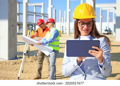 Beautiful female architect with tablet on construction site, behind her construction engineer and worker talking about the project, teamwork