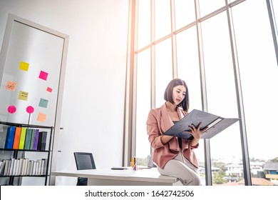 
Beautiful Female Asian Worker Holding A Folder File Of Documents And Information On Work Or Business Looking, Reading, Learning From The Documents, While Holding A Pen Working In  Office Environment