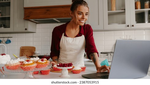 Beautiful female baker using laptop while making cakes and muffins on the kitchen - Powered by Shutterstock