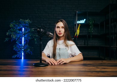 Beautiful female blogger sitting at a table with a microphone in the studio, recording blogs and looking at the camera with a smile on her face. - Shutterstock ID 2222007775