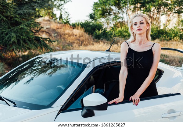 Beautiful female blonde driver in black dress by\
white car