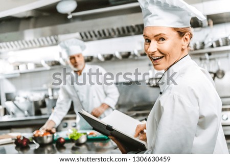 beautiful female chef in uniform holding recipe book and looking at camera in restaurant kitchen with colleague on background