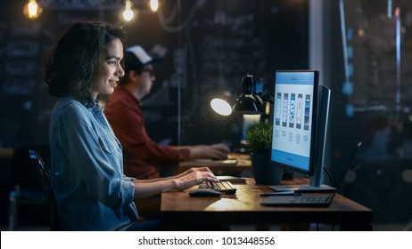 Beautiful Female Coder Works At Her Desktop On Her Peropnal Computer. Her Male Colleague Sits Next To Her. Evening Office Has Creative Lighting.