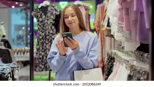 Beautiful Female Customer Shopping In Clothing Store, Using Smartphone, Browsing Online, Comparing On Internet, Choosing Stylish Clothes.