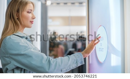Beautiful Female Customer Using Floor-Standing LCD Touch Display while Shopping in Clothing Store. She is Pressing Button to Start Using a Screen. People in Fashionable Shop.
