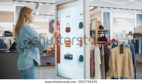 Beautiful Female Customer Using Floor-Standing LCD\
Touch Display while Shopping in Clothing Store. She is Choosing\
Stylish Bags, Picking Different Designs from Collection. People in\
Fashionable Shop.