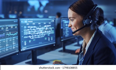 Beautiful Female Data Scientist Works On Personal Computer Wearing A Headset In Big Infrastructure Control And Monitoring Room. Woman Engineer In A Call Center Office Room With Colleagues.