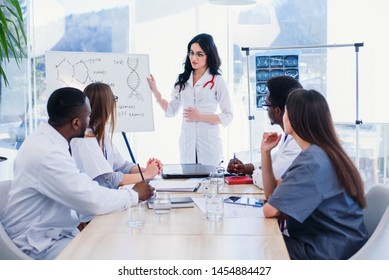 Beautiful Female Doctor Wearing White Coat And Eyeglasses Standing By Flipchart And Giving Presentation To Group Of Health Care Specialists. Medical Team Having A Meeting In Conference Room.