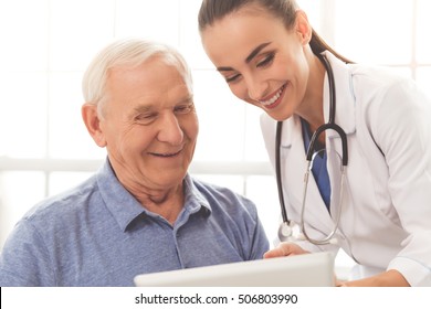 Beautiful Female Doctor In White Medical Coat Is Consulting Her Handsome Old Patient, Using A Digital Tablet And Smiling