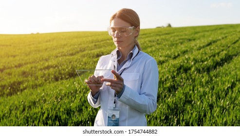 Beautiful female ecologist scientist in goggles standing in green field and working on glass transparent screen. Touchscreen of hi-tech. Scientist with smartphone of future in hands. 