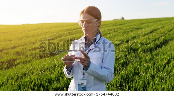 Beautiful\
female ecologist scientist in goggles standing in green field and\
working on glass transparent screen. Touchscreen of hi-tech.\
Scientist with smartphone of future in hands.\
