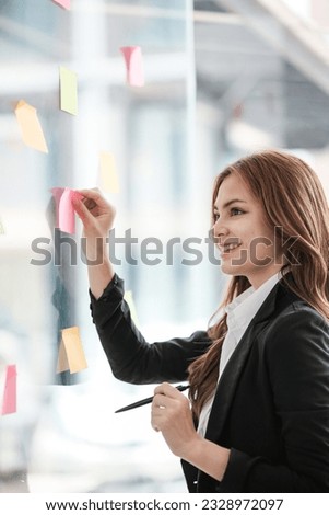 Beautiful female employee write down on colorful sticky notes manage list, concentrated biracial woman work on startup brainstorm collaborate plan on stickers on glass wall.