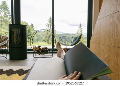 Beautiful female feet and book on background of  fireplace and big window in modern chalet. Young woman barefoot relaxing with book in comfortable home, cozy vacation. Leisure time - Shutterstock ID 1952568127