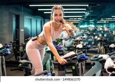 Beautiful Female Fitness Instructor With Headset And Microphone Pointing With Finger Straight On You While Doing Exercise On Indoor Stationary Bike During Cycling Training.