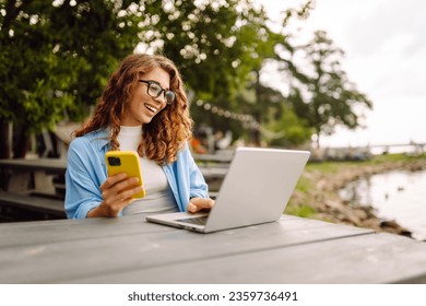 Beautiful female freelancer sitting on a bench at a table in a park outdoors, using a laptop and smartphone. Urban lifestyle. Business, blogging, freelancing.