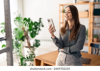 Beautiful female freelancer smiling and doing social media marketing over mobile phone while standing in living room. - Powered by Shutterstock