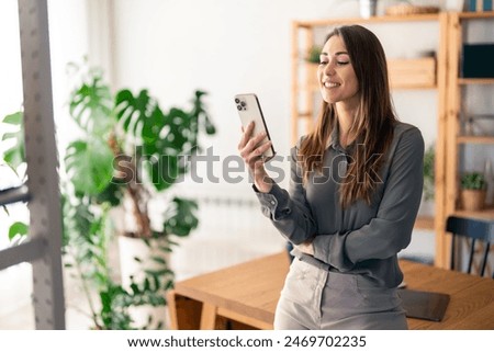 Beautiful female freelancer smiling and doing social media marketing over mobile phone while standing in living room.