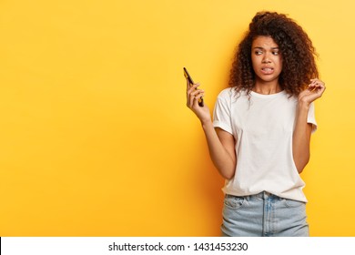 Beautiful female with frizzy Afro hairstyle, looks in displeasure at cell phone, receives call from former boyfriend, holds mobile phone on distance, ignores communication, afraids of something - Shutterstock ID 1431453230