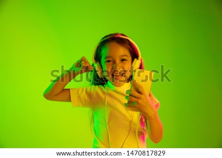 Beautiful female half-length portrait isolated on green backgroud in neon light. Young emotional girl. Human emotions, facial expression concept. Using smartphone for vlog, selfie, chating, gaming.