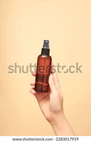 Beautiful female hand holding an empty bottle without label on beige background. Mockup for cosmetic spray bottle such as mineral spray or hair spray. Front view, space for design.