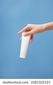 Beautiful female hand holding a tube in white color on blue background. Empty label for product mockup. View from front