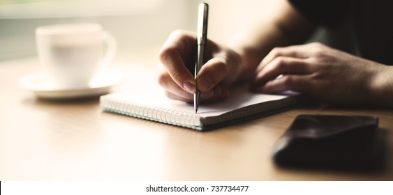 Beautiful Female hands with phone and cup of coffee write pen in a notebook of tasks and goals to work on a wooden table - Powered by Shutterstock