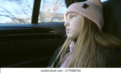 Beautiful Female Kid With Long Blonde Hair Looks Outside Through The Window In The Backseat Of A Moving Car. Small Sad Girl Rides On Modern SUV And Watches At Nature Through The Glass Of Auto. Slow Mo