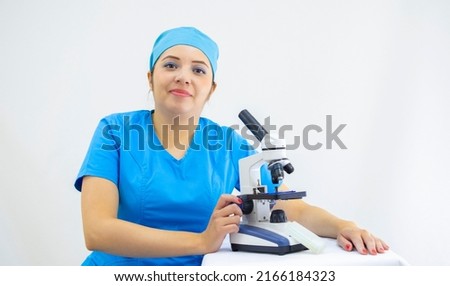 beautiful female lab technician wearing uniform and blue surgical cap, analyzing samples with the microscope, on white background