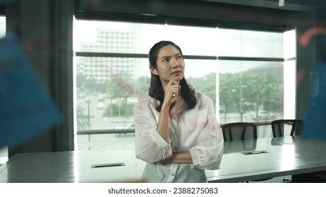 Beautiful female leader thinking about start up project in front of glass board. Smiling young project manager looking at colorful sticky notes with show creativity marketing strategy. Manipulator.