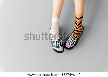 Beautiful female legs in missmatched teen socks wearing fashionable rubber slippers with inscription why not on gray background. Elegant stylish trendy and voguish footwear for fashionable ladies