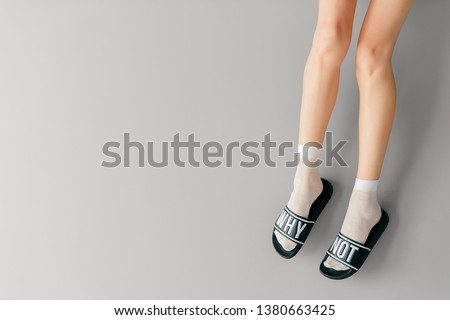 Beautiful female legs in white mesh teen socks wearing fashionable rubber slippers with inscroption why noton gray background. Elegant stylish trendy leg-wear . Voguish legwear for fashionable ladies