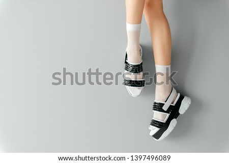 Beautiful female legs in white mesh trendy socks wearing fashionable black white high wedge leather sandals. Womens modern fashion footwear. Rebel girl in high sole stylish shoes on gray background