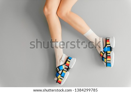 Beautiful female legs in white trendy socks posing in fashionable colorful high wedge leather sandals on gray background. Womens modern voguish footwear. Girl wearing high sole summer massive shoes.